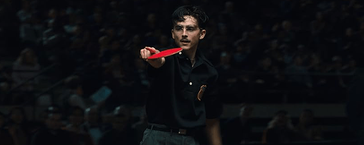 Timothée Chalamet wearing glasses and holding a table tennis racket points towards someone just outside of the frame