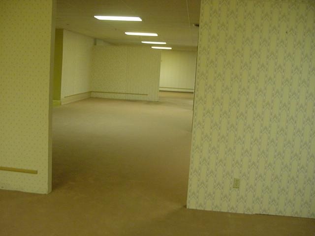A room with yellow wallpaper, seemingly empty with a strange pattern on the wallpaper, a carpetted floor that appears well worn and several overhead lights 